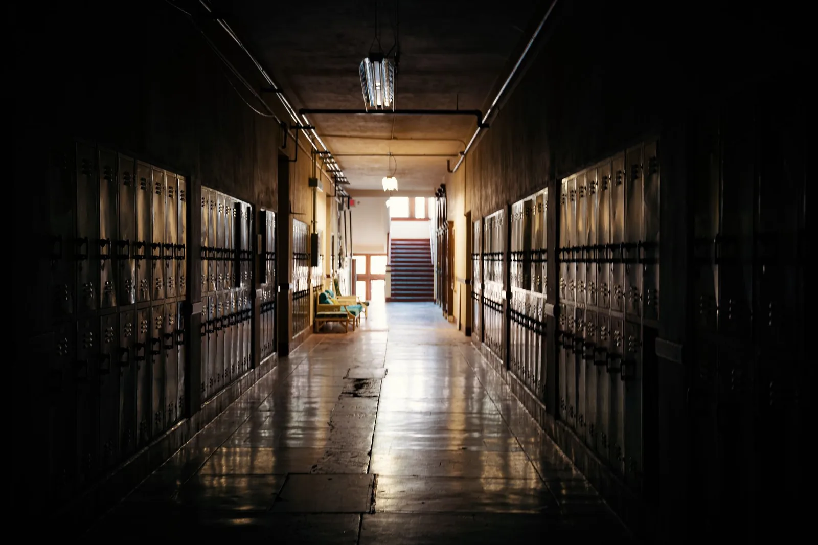 An empty school hallway lined with metal lockers, lit by daylight from a stairwell at the far end.