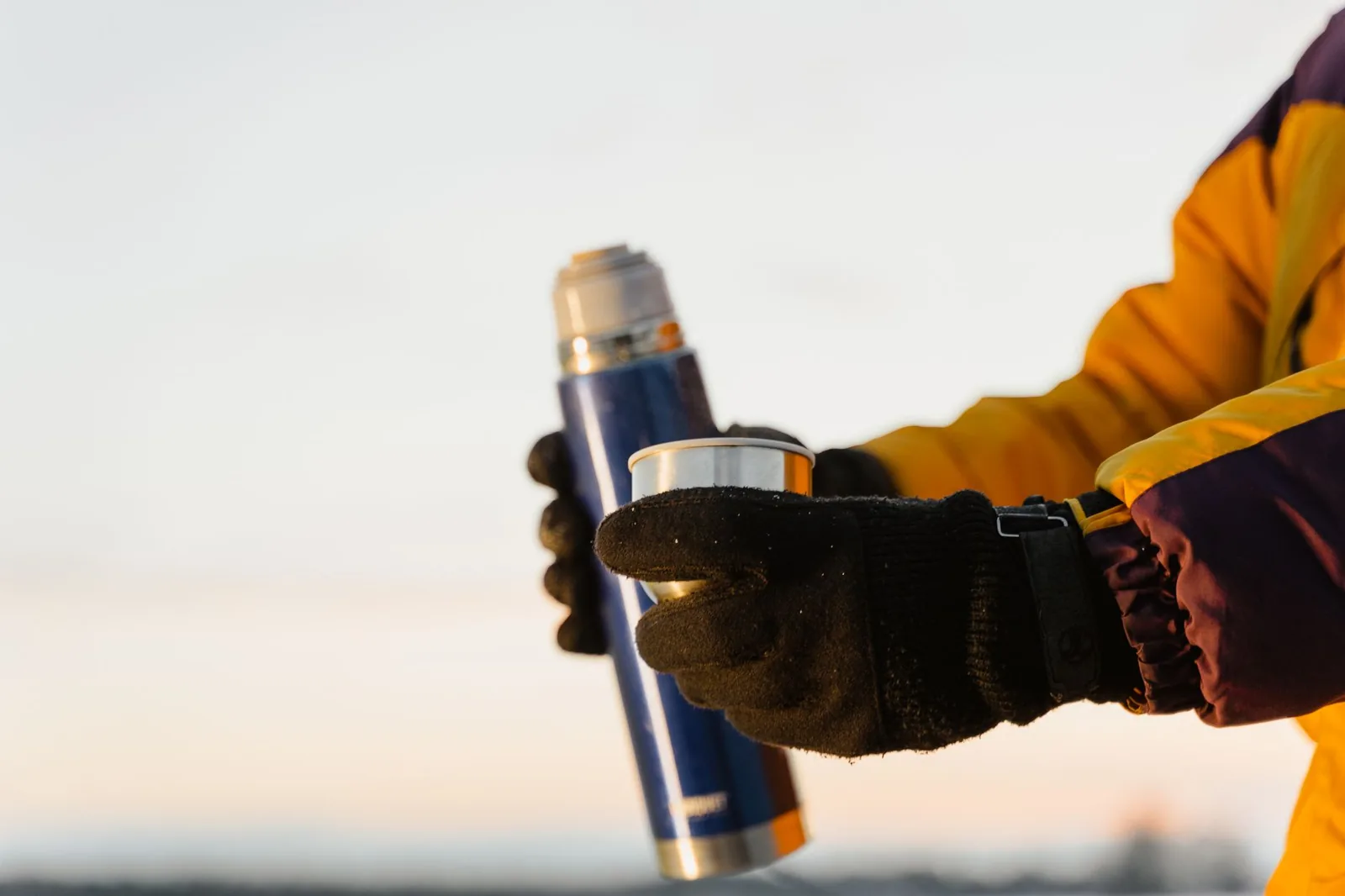 Gloved hands holding a thermos and cup outdoors